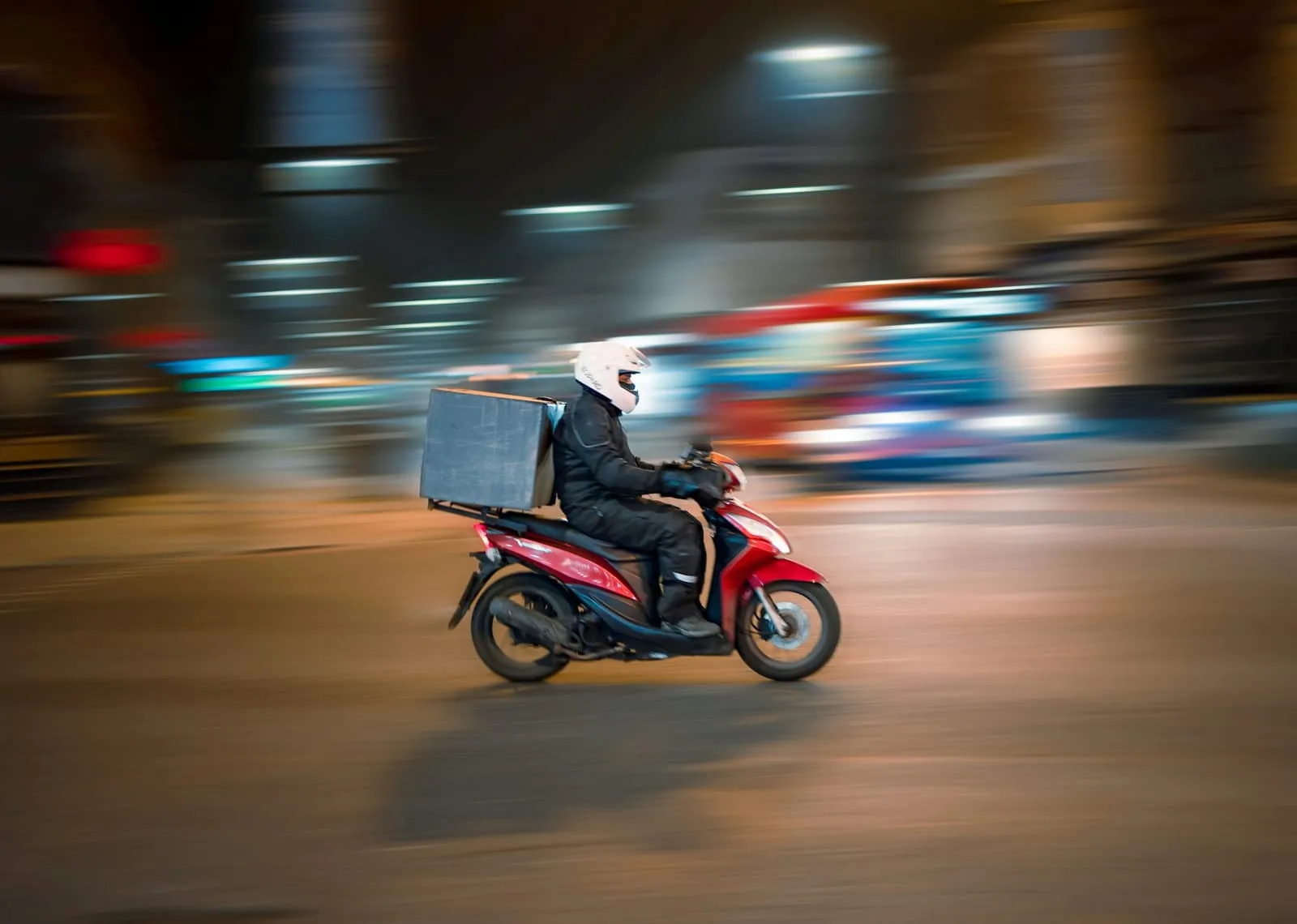 A delivery rider on a red scooter riding through a city at night